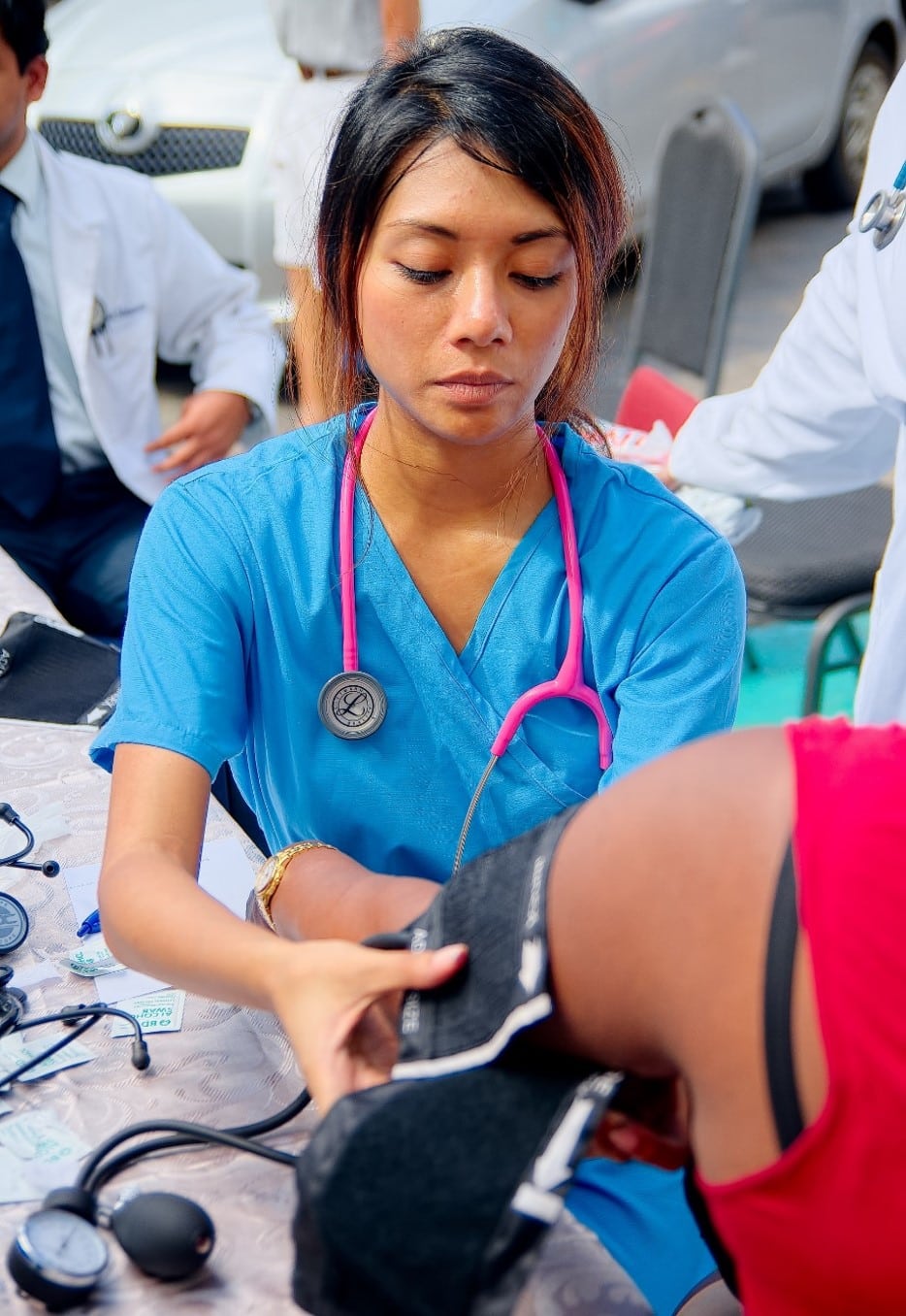 The image shows a medical professional, likely a student or nurse, using a blood pressure cuff to check a patient's blood pressure. The setting appears to be a clinical or community health event, possibly organized by a medical school like All Saints University (ASU), given the context of your previous question. Other individuals in medical attire are visible in the background, suggesting a group effort, perhaps a health outreach program.