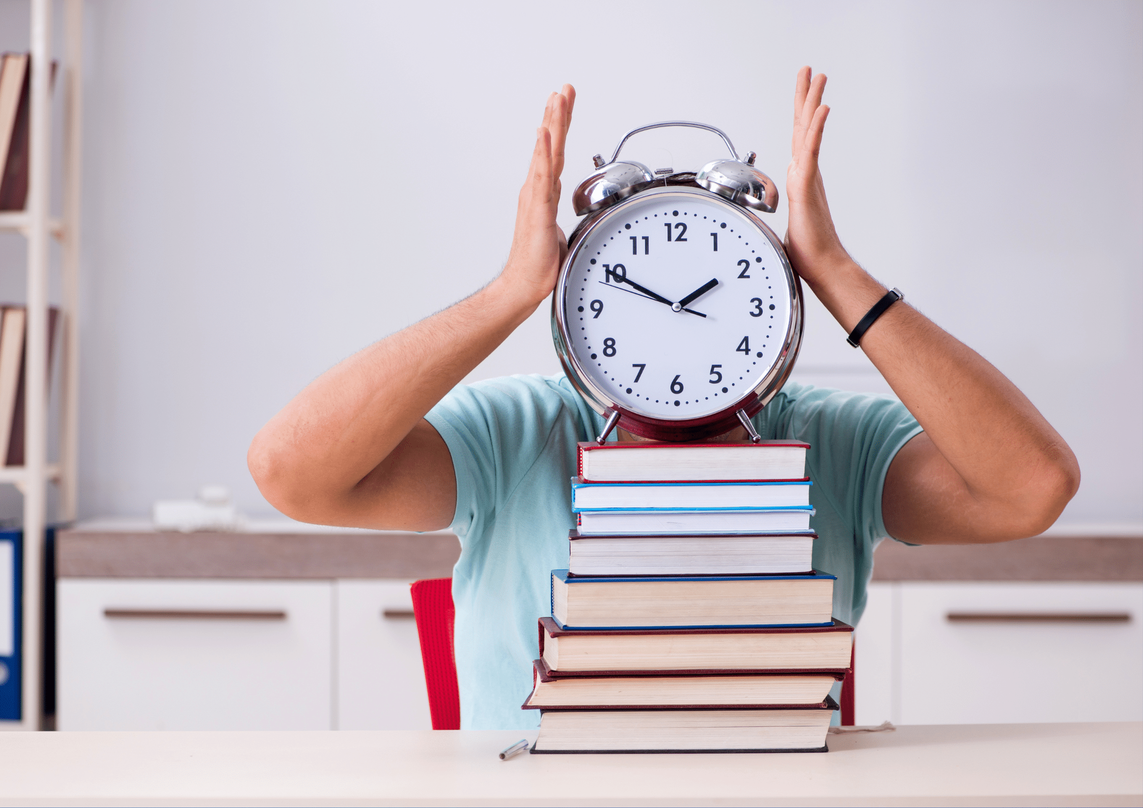 A person wearing a light blue shirt is sitting at a desk with a stack of colorful books in front of them. The person is holding a large, old-fashioned alarm clock with both hands, covering their face, with the clock showing a time close to 12. The background features a modern room with white cabinets, a shelf with books, and a red chair.